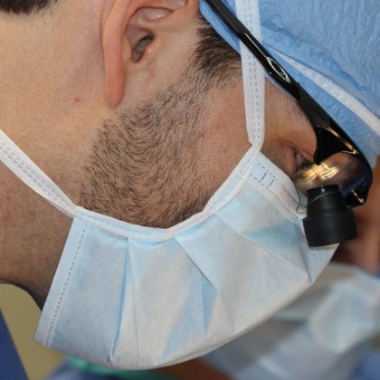 A veterinarian currently performing surgery. Vet is dressed in scrubs and wearing a mask, cap and glasses